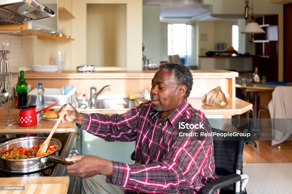 Disabled Afro-American Senior Man in Wheelchair Preparing Food Disabled Afro-American Person is Enjoying in Preparation of Delicious Lunch. A Senior African is Cooking a Dinner in Domestic Kitchen. Cooking Stock Photo