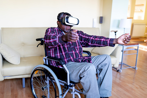 Cheerful Disabled Senior Afro-American Man, in a Wheelchair, is Gesturing and Smiling, While Wearing the Virtual Reality Goggles at Home in the Livingroom.