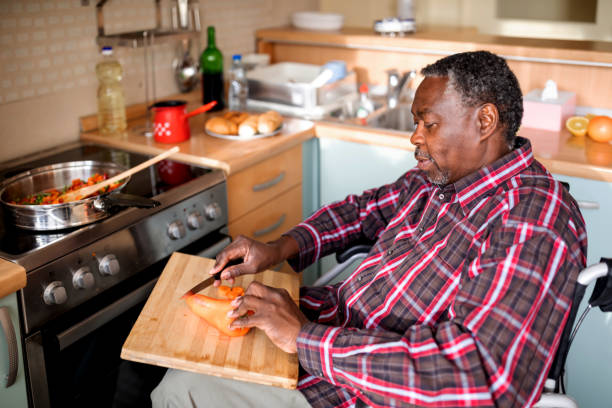 istock Senior Man with mobility handicap slicing red pepper using sharp knife in kitchen. Disabled paralyzed handicapped man with walking disability integrating after an accident. 1286892163
