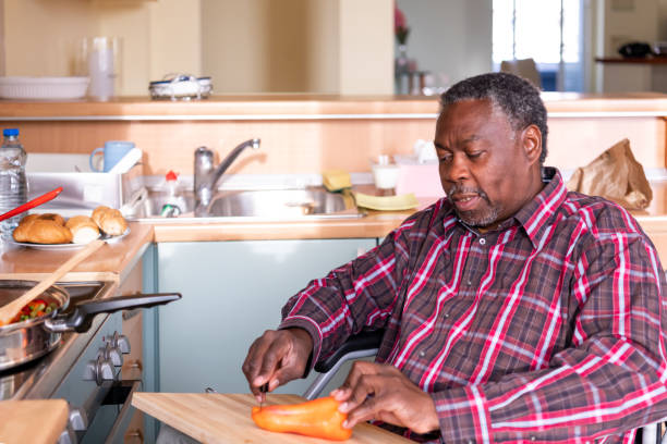 istock Senior Man with mobility handicap slicing red pepper using sharp knife in kitchen. Disabled paralyzed handicapped man with walking disability integrating after an accident. 1286892167