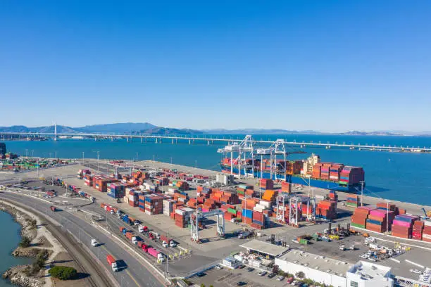An aerial view of the Port of Oakland. Colorful cargo containers fill the yard and are off loaded from cargo ships. Bay Bridge in the background.