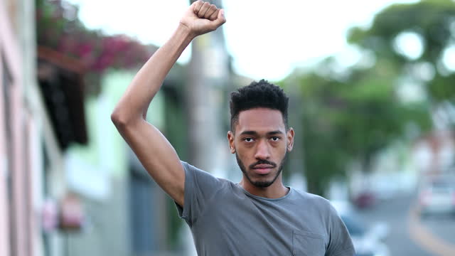 Individual young black man raising fist in air staring camera in protest. Rebel African mixed race ethnicity