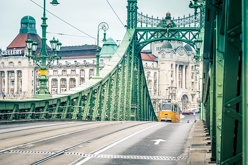 The famous yellow tram passing over the Liberty bridge in Budapest. Old yellow traim service is still running during the covid-19 situation. Famous Gellert Spa hotel in the background.
