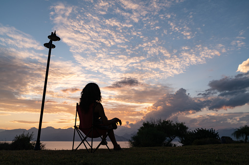 Silhouette of woman sitting on camping chair by sea during sunset. Sky has high clouds and is orange. Shot in outdoor daylight with a full frame mirrorless camera.