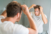 Hair loss man looking in bathroom mirror putting wax touching his hair styling or checking for hair loss problem. Male problem of losing hairs