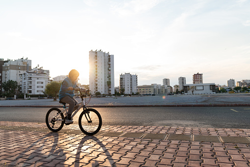 Photo of 7,5 years old elementary schoolboy riding bike on pavement in public park during sunset. City buildings are seen on the background. Shot with a full frame mirrorless camera.
