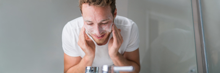 Man washing face banner panorama. Person cleansing with facial cleanser face wash soap in bathroom sink at home.