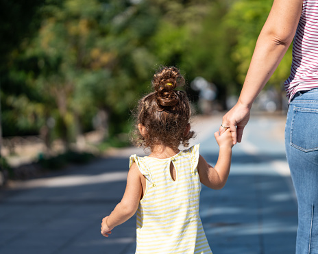 Rear view of 2,5 years old girl holding mother's hand and walking in public park. She is wearing a yellow dress. Shot in outdoor with a full frame mirrorless camera.