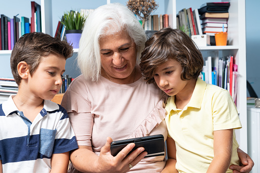 Senior grandmother and two elementary school boys using smartphone in front of bookshelf in living room. Senior woman has white hair and sitting in the middle of two grandsons. Shot with a full frame mirrorless camera.