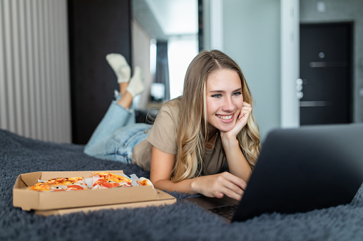 Young woman lying on a bed in bed at home while eating pizza and using laptop
