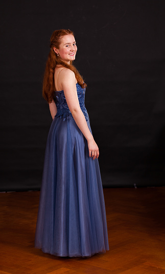 Dressed in a blue classical dress, a 17 year old natural red haired teenage girl is standing full length against a dark background.