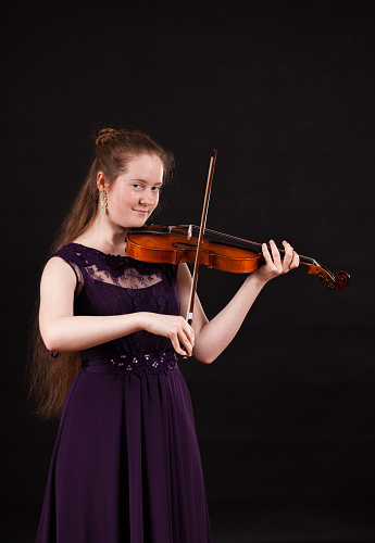 15 year old girl in purple classic dress looks at camera while playing classical music on the violin. Copy space provided.