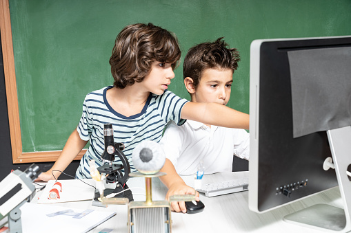 Two elementary school students building robotics in STEM class. A green chalkboard is seen on the background. A small humanoid robot is seen on foreground.They are looking at the computer.  Shot indoor with a full frame mirrorless camera.
