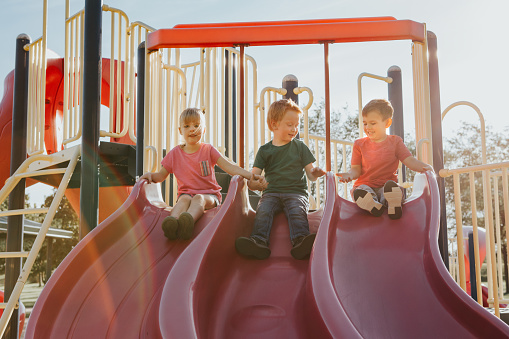 Active happy Caucasian children sliding on playground schoolyard outdoor on summer sunny day. Kid friends boys girl having fun. Seasonal kids activity outside. Authentic childhood lifestyle concept.