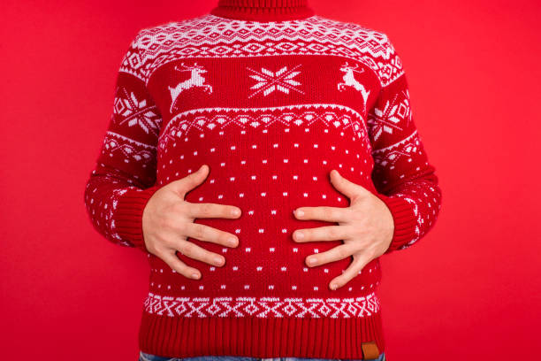Holidays overeating concept. Close up photo of men in red winter sweater with deers holding his big tummy isolated on red background Holidays overeating concept. Close up photo of men in red winter sweater with deers holding his big tummy isolated on red background over eating stock pictures, royalty-free photos & images