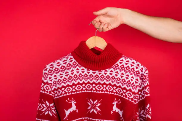 Photo of Ugly Christmas sweater party concept. Close up photo of men holding hanger with red winter sweater with deers  isolated on red background