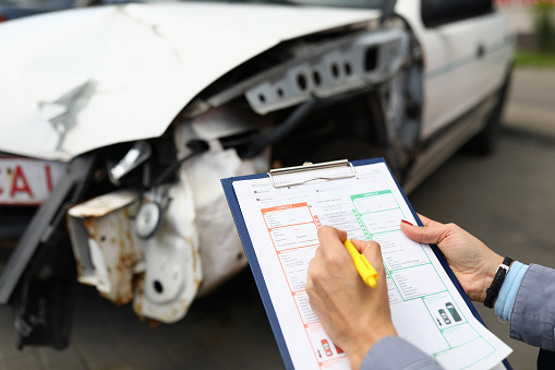 Insurance agent holds clipboard and ballpoint pen closeup and wrecked car in background. Vehicle insurance concept.