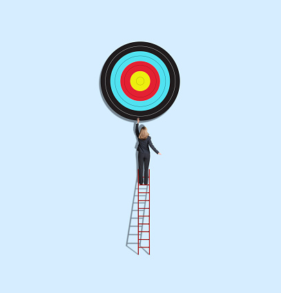 A businesswoman stands on one of the higher rungs of a red ladder as she stretches to reach for a large target above her.