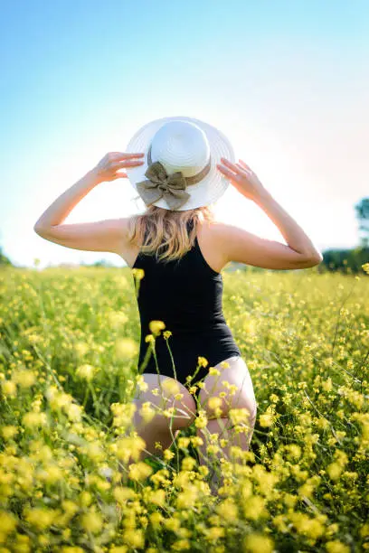 Blonde girl in a black bodysuit with a white hat walks in a rapeseed field, having fun, view from the back