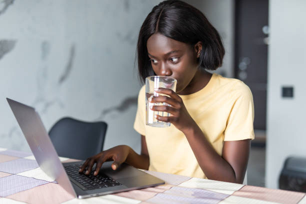 istock Beautiful young black woman using laptop computer on kitchen while drink water 1286990740