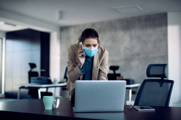 istock Entrepreneur with face mask using laptop and communicating over mobile phone at her office 1286990916