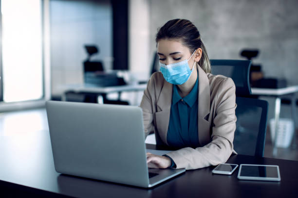istock Businesswoman typing an e-mail on laptop while wearing protective face mask in the office. 1286992080