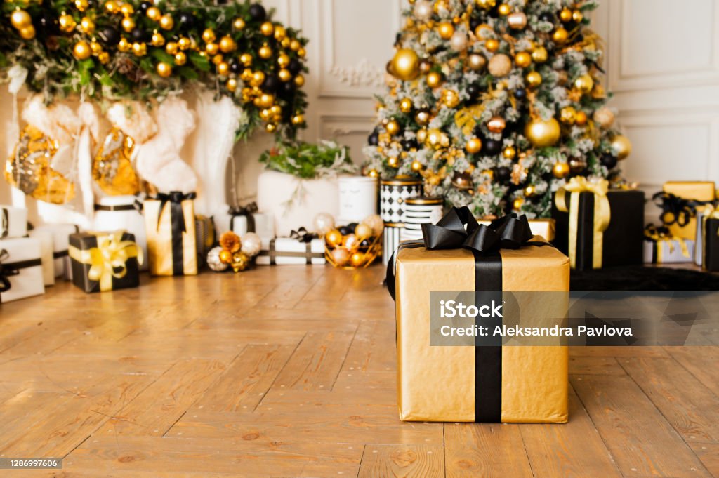 gold gift box with black ribbon on wooden floor in room with Christmas decor in gold and black colors : Christmas tree with balls and garlands and many Packed gifts under it, selective focus Celebration Stock Photo