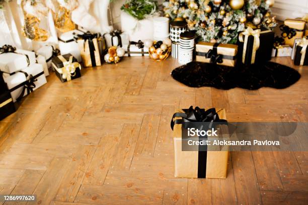 Gold Gift Box With Black Ribbon On Wooden Floor In Room With Christmas Decor In Gold And Black Colors Christmas Tree With Balls And Garlands And Many Packed Gifts Under It Selective Focus Stock Photo - Download Image Now