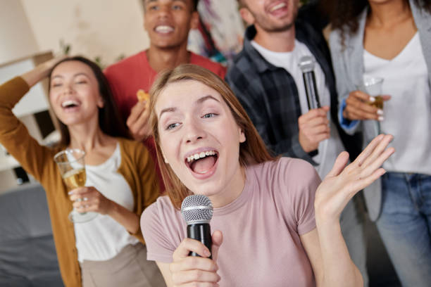 istock Sharing Joy. Beautiful young girl holding microphone and singing while playing karaoke with best friends at home, having great time together 1287000230
