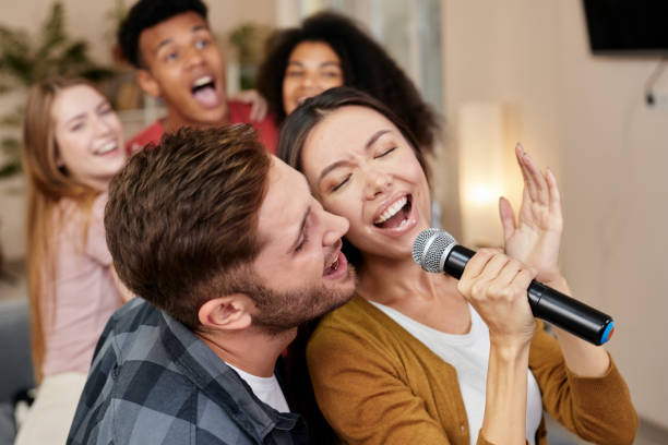 istock Sing together. Young excited couple or friends holding microphone and singing together while playing karaoke with friends at home. Group of people having fun at karaoke home party 1287000331