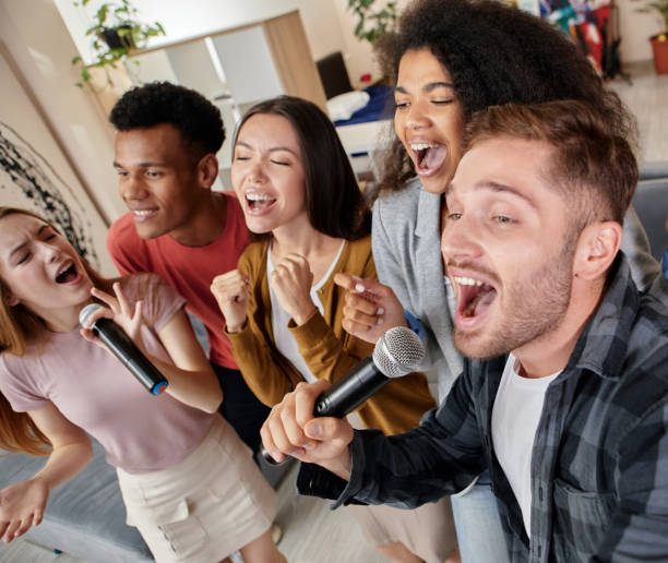 istock Face the song. Enthusiastic multicultured friends singing with microphone while playing karaoke at home, standing in the modern apartment 1287000446