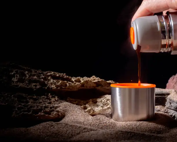 Hand pouring coffee from a stainless steel thermos into a metal mug. Keep the drink hot in a steel drum to enjoy a hot cup of coffee on a cold night.
