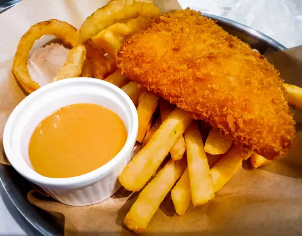 Deep fried fish made into fish and chips menu served with french fries, onion rings and orange-color dipping sauce in the metallic tray on the table in some fast food restaurant.