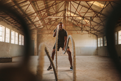 Tough man working out in cross training gym made inside old factory. Fitness man exercising with battle rope in abandoned warehouse.