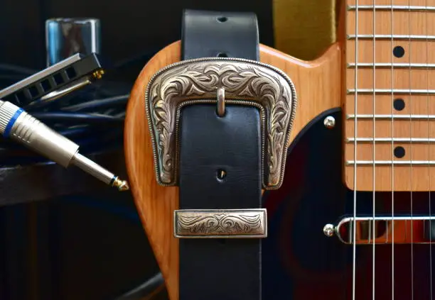 Photo of Electric guitar and leather strap with silver buckle. Close-up with glass slide, blues harp and cable jack.