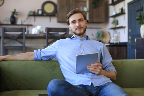 istock Smiling handsome man holding and using tablet with social networks at home, happy freelancer making online shopping, sitting on cozy sofa at living room. 1287027886