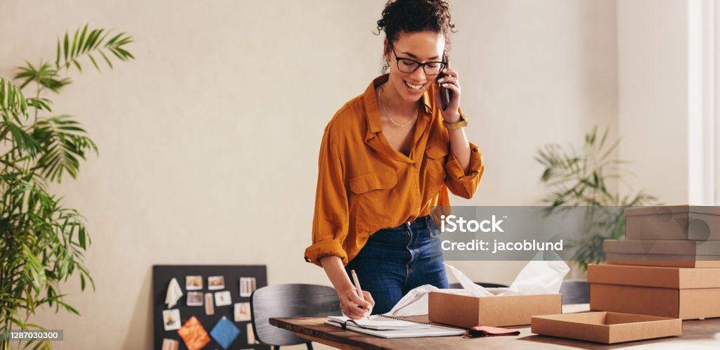 Drop shipping business owner confirming the order on phone Drop shipping business owner talking on mobile phone and taking order. Female entrepreneur working at home office confirming the order on phone. Small Business Stock Photo