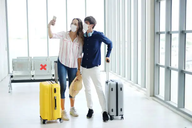 Photo of Young Caucasian traveler couple love wear mask for prevention for covid-19 virus and selfie with smartphone together in the airport. Healthcare and technology for Honeymoon trip on vacation Concept.