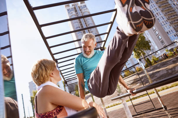 istock Smiling mature man in a great shape doing push ups on parallel bars and talking with his wife. Healthy mature couple doing sport together in the morning outdoors 1287040252