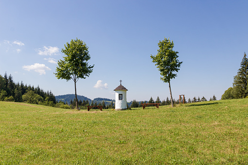 Small white round chapel with two wooden benches between two young leaf trees. Green meadow and place for relaxing. Summer day in Czech countryside.