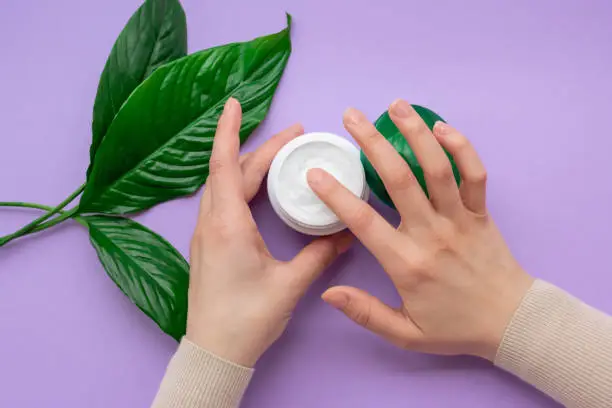 Girls hand is nearby jar with white soft hand and body cream on purple background with large green leaves.Concept of eco cosmetic.Girl is moisturizing her hand by cream as beauty procedure at home.