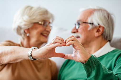 Happy seniors posing with holding hands and smiling. Relationships, love and old people concept - close up of senior couple showing hand heart gesture