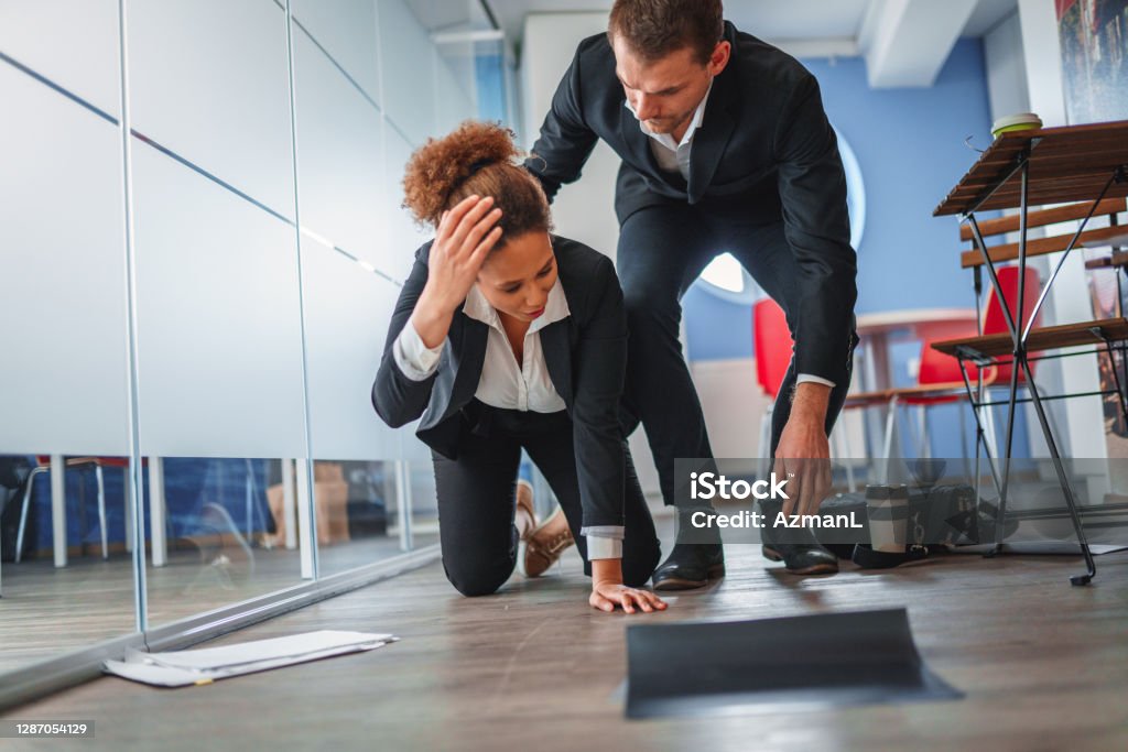 African-American Businesswoman Falling and Dropping Papers in an Office African-American businesswoman falling and dropping paper on the floor in an office. Caucasian businessman is helping her. Misfortune Stock Photo