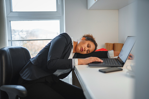 African-American businesswoman falling a sleep in an office while using laptop. She is leaning on the table.