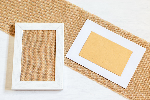 Zero waste ecological blank frame for card design. White wooden picture frame with burlap tuxtered fabric, craft envelope with copy space. Handmade mock up things, DIY concepts.