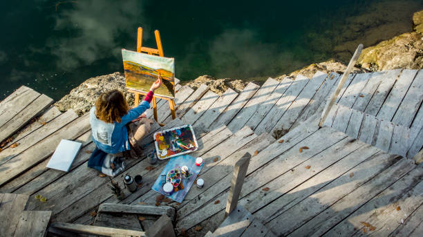 a drone shot of a young artist painting in nature by a beautiful lake - a young beautiful artist inspired by autumn nature creates her work of art - easel art paint artists canvas imagens e fotografias de stock