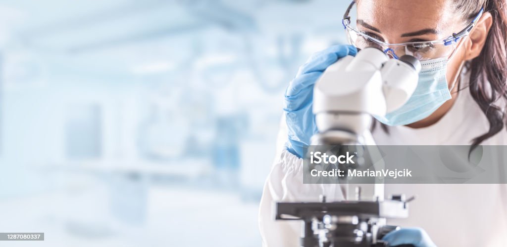 Die Labortechnikerin in Schutzbrille, Handschuhen und Gesichtsmaske sitzt neben einem Mikroskop im Labor. - Lizenzfrei Labor Stock-Foto