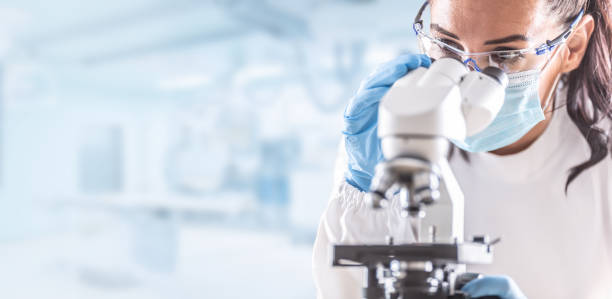 Female lab technician in protective glasses, gloves and face mask sits next to a microscope in laboratory.