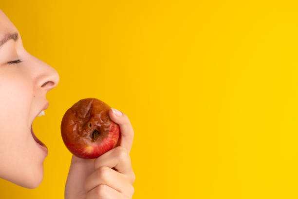 The girl bites a rotten apple with a worm on a yellow background. Expired products, junk food The girl bites a rotten apple with a worm on a yellow background. Expired products, junk food. rotting apple fruit wrinkled stock pictures, royalty-free photos & images