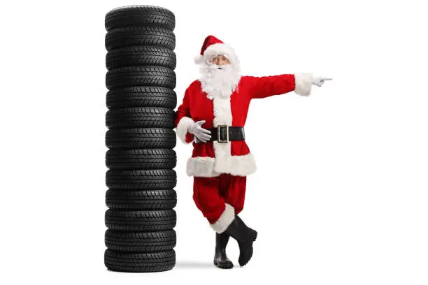Full length portrait of santa claus leaning on a pile of tires and pointing to the side isolated on white background